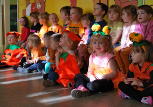 Dzieci siedzą na podłodze, uśmiechają się, są ubrane w pomarańczowe stroje